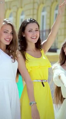 Canlı üç kız, renkli yaz kıyafetleri içinde güneşli bir caddede yürürken coşkulu bir şekilde alışveriş çantalarını sallıyorlar. Bulaşıcı gülüşleri ve ışıl ışıl gülüşleri canlı bir dostluk ve kutlama ortamı yaratıyor.