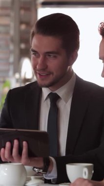 Üç iyi giyimli adam şık bir kafede lezzetli bir öğle yemeğinde stratejileri tartışırken gülümsemeleri ve hareketleriyle coşku saçıyorlar. Sıcak kahve ve parlak bir tabletle birlikte davetkar atmosfer, dinamik takım çalışmalarını ön plana çıkarıyor.