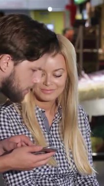 Çekici ve genç bir çift, işlerine tamamen dalmış bir şekilde, canlı bir süpermarket sahnesinin ortasında market fiyatlarını hesaplıyor. Sakallı adam akıllı telefonuna dikkatle odaklanırken sarışın kadın renkli raflarla çevrili neşeyle ışıldıyor.