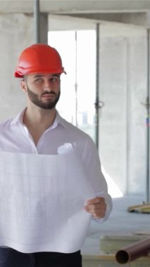 Canlı turuncu şapkalı genç, sakallı bir inşaat mühendisi inşaat halindeki binayı inceliyor, elinde kendine güvenen bir gülümsemeyle ayrıntılı planlar tutuyor. Ham beton ve yüksek iskele ile çevrelenmiş,