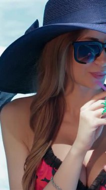 Parlak güneş şapkalı, kahverengi saçlı beyaz bir kadın havuz kenarında renkli bir kokteylin tadını çıkarıyor. Güneş ışınlarını yansıtan şık güneş gözlükleriyle, içkisini neşeyle yudumluyor, yaz tatili ve tropik havanın esansını yayıyor.