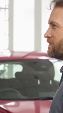 Zevkli, sakallı bir adam, lüks bir galeride çarpan kırmızı bir otomobilin yanında duran arabasının anahtarlarını tutarken mutluluk saçar. Bu heyecan verici an, yeni bir mal satın almanın sevincini ve gururunu yansıtıyor.