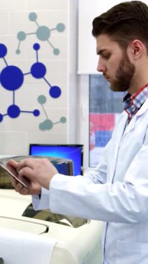 Kendini adamış beyaz laboratuvar önlüklü genç beyaz bir bilim adamı titizlikle bir bilgisayardan son teknoloji laboratuvar ekipmanlarıyla çevrili parlak bir tablete veri aktarıyor. Onun düşünceli ifadesi ve düzgün taranmış saçı bir bağlılığı yansıtıyor.