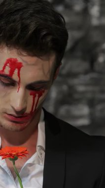 Yüzünde kan kırmızısı çizgileri olan parlak siyah takım elbiseli bir erkek vampir, ürkütücü bir şekilde turuncu bir çiçeğin yapraklarını koparır, gölgeli tuğla bir duvara yaslanır. Bu büyüleyici sahne, Cadılar Bayramı 'nın tüyler ürpertici ruhunu çağrıştırıyor.