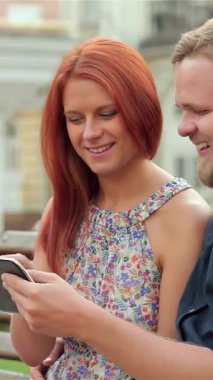 Göz alıcı bir çift, canlı bir şehir parkında gülümseme ve kahkaha atarken tabletleri tarafından büyülenen, kırsal bir ahşap bankta yakın bir şekilde oturuyor. Altın güneş ışığı yüzlerini yıkıyor ve yaz kaçamaklarının neşeli atmosferini güçlendiriyor.