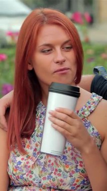 Neşeli bir çift, güneşli bir park bankında oturuyor, canlı güllerle çevrili iken, kahvelerini yudumluyorlar. Neşeli şakalaşmaları ve sıcak gülümsemeleri bu açık havada dostluk ve bağlılık için davetkar bir ortam yaratır.