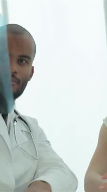 Parlak, modern bir klinikte, beyaz önlüklü, odaklanmış bir erkek doktor, dikkatli kadın hastasıyla ciddi bir konuşma yaparken göğüs röntgeni çeker. Atmosfer profesyonellik ve özen gösteriyor.