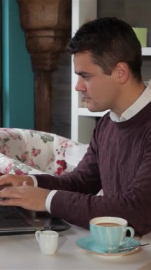 Şık bir beyaz adam dizüstü bilgisayarında yazarken neşeyle ışıldıyor şık bir kafede zengin bir fincan kahvenin tadını çıkarıyor. Çiçekli koltuklar ve güneşli bir pencereyle çevrelenmiş sahne, sıcaklığı ve yaratıcılığı yayarak üretkenliği gevşek bir titreşim ile harmanlıyor.