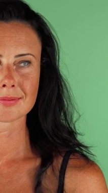 Çarpıcı, olgun bir kadın, dalgalı, koyu saçlı, hoş bir şaşkınlık ifadesi, eli zarif bir şekilde ağzını canlı bir kromakey yeşil arka planına karşı kapatıyor. Bu büyüleyici görüntü güven ve cazibe yayıyor, izleyicileri davet ediyor.