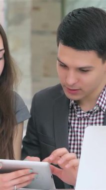 Genç, çekici bir iş adamı ve kadın, şık bir tablet üzerindeki verileri incelerken canlı bir tartışmaya girişiyor ve kadın kameraya içten bir şekilde gülümsüyor. Keskin profesyonel kıyafetleri ve odaklanmış ifadeleri takım çalışması atmosferi yayıyor.