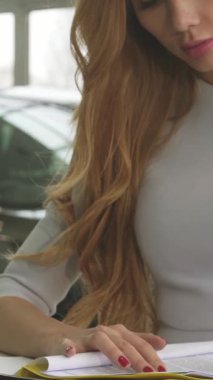 Uzun, dalgalı kızıl saçlı şık bir kadın, parlak, modern araçlarla çevrili çağdaş bir araba galerisinde evrak imzalamakla derinden meşgul. Bu dinamik sahne, yeni bir araba almanın heyecanını özetliyor.