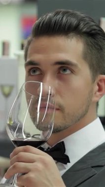 Zarif bir yelek ve papyonlu şık genç bir erkek şarap garsonu zarif bir şekilde koyu kırmızı şarap bardağı tutuyor ve odaklanmış bir takdir ifadesi ile karışık aromalarını içine çekiyor. Şık düzenlenmiş şarap şişelerinin arka planına karşı bu sahne...