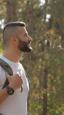 Sakallı bir erkek yürüyüşçü elini sevinçle gökyüzüne doğru tutar ve güzel esmer kız arkadaşıyla keyifli bir anı paylaşır. Altın güneş ışığıyla yıkanan çift, güneşle ıslanmış ormandaki görkemli ağaçların arasında duruyor.