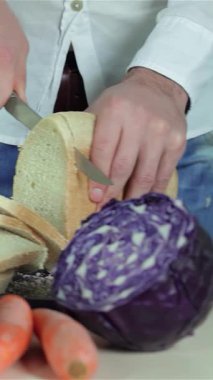 Sıcak bir mutfakta, bir çift el, mor lahana ve parlak turuncu havuç gibi renkli sebzelerle çevrili bir somun taze ekmeği ustalıkla dilimliyor. Sahne sıcaklığı ve yaratıcılığı yansıtıyor, izleyicileri mutfağa davet ediyor.