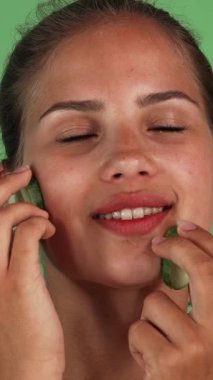 Göz kamaştırıcı genç bir kadın yüzüne serin salatalık dilimleriyle neşeli bir şekilde masaj yapar, canlı yeşil bir krom arka plana tazelik saçar. Bu canlandırıcı cilt bakım ve tazeleme ayini, izleyicileri davet ederek özgüvenin özünü yakalar.