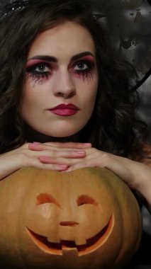 Büyüleyici bir büyücü, gotik kıyafetlerle süslenmiş, oyulmuş bir balkabağı ile zarifçe poz veriyor, sırıtan yüzü gölgeli bir tuğla zemin üzerinde parlıyor. Çarpıcı makyajı ve dalgalı saçları büyüleyici Cadılar Bayramı atmosferini güçlendiriyor.