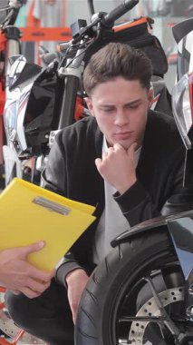 Canlı bir motosiklet galerisinde, şık siyah ceketli genç bir adam zarif siyah bir spor motosikleti inceliyor. Kareli gömlekli bir danışman ise etkileyici özelliklerini coşkuyla paylaşıyor. Hava heyecanla dolu.