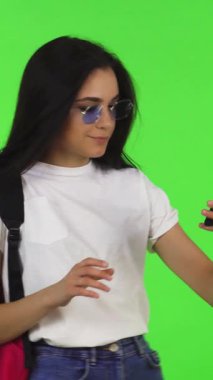 Uzun, gösterişli saçları ve şık güneş gözlükleriyle şık genç bir kadın canlı yeşil bir kromakey arka planına kurulmuş akıllı telefonuyla neşe içinde selfie çekiyor. Bu enerjik sahne, çağdaş öğrenci hayatının özünü yansıtıyor.