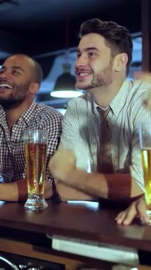 İki heyecanlı arkadaş, şık şık, şık, sıradan kıyafetler giyiyorlar, tutkulu bir şekilde tezahürat yapıyorlar, soğuk bardaklarını kaldırırken, hareketli bir spor barının heyecan verici atmosferine dalıyorlar. Sahne kahkahalarla ve arkadaşlıkla dolu.