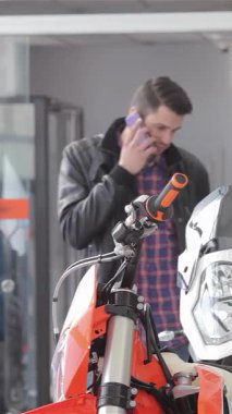 Şık, esmer bir adam şık turuncu bir motosikletin yanında duruyor, canlı bir galeride derin bir telefon görüşmesi yapıyor. Kareli ceketi dinamik atmosferi tamamlıyor. Motosiklet ve uzmanlık tutkusunu gösteriyor.