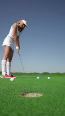 Şık beyaz elbiseli çarpıcı kızıl saçlı bir kadın, üç golf topuna verimli, canlı yeşil bir sahada ustalıkla vururken şiddetle konsantre oluyor. Sahne kararlılık ve kesinlik saçıyor, toplar deliğin kenarında sallanıyor.