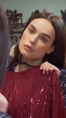 Sevimli genç bir kadın aynanın önünde duruyor, zarif bir şekilde iki zarif elbiseyi tutuyor, moda bir butikte şık seçeneklerini değerlendirirken düşüncelere dalmış. Giysilerin zengin dokuları ve canlı renkleri onun kusursuzluğunu yansıtıyor.