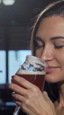 Uzun, dalgalı saçları olan çekici bir kadın ferahlatıcı bir zanaat birasından zevk alıyor. Işıl ışıl gülüşü, düşünceli bir şekilde barın canlı atmosferine bakarken saf mutluluğu yansıtıyor. Kırsal ahşap bar ve yumuşak, ılık ışıklandırma davetkar bir zemin oluşturur.