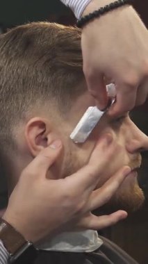 Bu yakın plan çekimde şık, bakımlı sakallı genç bir adam, profesyonel bir berber titizlikle yüzünü jiletle tıraş ediyor. Klasik berber dükkanı atmosferi cazibe saçıyor, geleneksel tımar sanatını ön plana çıkarıyor ve...