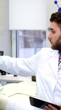 Odaklanmış, şık sakallı beyaz bir adam son teknoloji ürünü bir laboratuardaki düzgün bir bilgisayar ve tablet üzerindeki verileri titizlikle analiz ediyor. Onun yanında, gri saçlı kıdemli bir laboratuvar çalışanı düşünceli bir şekilde başını sallıyor ve canlı bir işbirliği sahnesi sergiliyor.