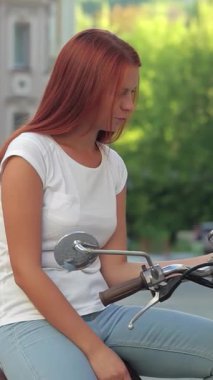 Dalgalı kızıl saçlı neşeli bir genç kız şık scooterına biniyor, resim gibi bir Avrupa şehrinin canlı atmosferini kucaklıyor. Güneşli sahne bir macera ve özgürlük hissi yayıyor, izleyicileri yazın zevkine davet ediyor.