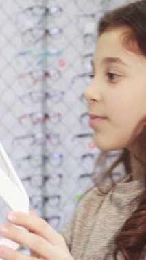 Dalgalı kahverengi saçlı neşeli küçük bir kız, göz kamaştırıcı bir gözlük mağazasında göz kamaştırıcı bir güneş gözlüğü seçimi yaparken heyecan saçıyor. Güneş gözlüklerinin canlı renkleri ve şık tasarımları hayal gücünü ateşliyor.