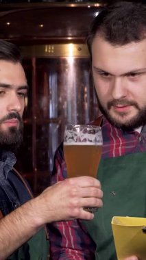 İki kendini adamış erkek bira üreticisi, biri şık sakallı, canlı altın biralarını ve zengin koyu biralarını dikkatle analiz ediyor. Pırıl pırıl paslanmaz çelik ekipmanlarla çevrelenmiş bu canlı tartışma sanatsal ve takım çalışmasını yansıtıyor.