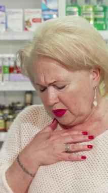 Acılı bir yüz ifadesi ve eli alnında yaşlı bir kadın, canlı bir eczane koridorunda çaresizce ilaç arıyor. Renkli sağlık ürünleri ile çevrelenmiş, hastalıktan kurtulma mücadelesi,