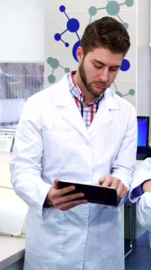 Gösterişli, yüksek teknolojili bir laboratuvarda odaklanmış esmer bir stajyer, gri saçlı kıdemli meslektaşıyla monitörden görüşlerini paylaşıyor. Canlı atmosfer gelişmiş laboratuvar ekipmanları ve dijital tablet ile zenginleştiriliyor.