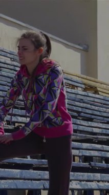 Kararlı genç bir kız, hareketli bir stadyumun hareketli tribünlerinde bacaklarını ve ayaklarını uzatarak odak ve atletizm saçıyor. Renkli bir geometrik kapüşonlu ve rahat atletik pantolonla hazırlanırken kendini adama ruhunu somutlaştırıyor.