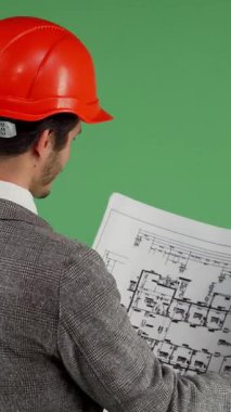 Çarpıcı turuncu şapkalı neşeli sakallı bir adam ve özel dikim bir takım elbise detaylı mimari planları inceliyor, özgüven ve profesyonellik yayıyor. Sıcak gülüşü canlı yeşil bir arka plana karşı davetkar bir atmosfer yaratıyor.