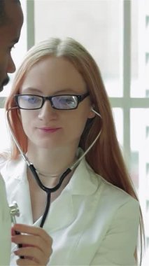 Güneşli bir klinikte, şefkatli bir bayan doktor steteskopunu kullanarak genç bir adamın kalp atışlarını dikkatle inceliyor, profesyonellik ve bakım yayıyor. Nazik gülüşü ve odaklanmış bakışları güven veren bir çevreyi teşvik ediyor.