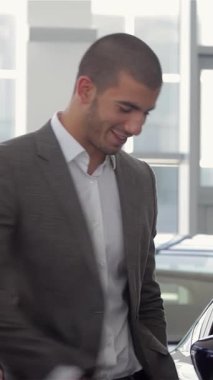 Parlak, modern bir araba galerisinde tutkulu bir araba satıcısı şık giyimli genç bir adama enerjik bir şekilde yeni bir araba gösteriyor. Satış elemanı hareketli hareketlerle arabaların etkileyici özelliklerini sergiledikçe hava heyecanla doluyor.