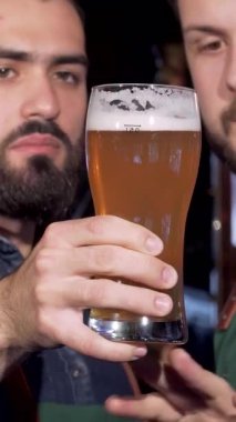 İki yetenekli bira imalatçısı, biri kot gömlekli, diğeri ekose gömlekli, bir bardak altın zanaat birasını dikkatle inceleyerek, kaliteli biraya olan bağlılıklarını vurguluyorlar. Bira fabrikasının sıcak, davetkar atmosferi, parıldayan bakır ekipmanları ve