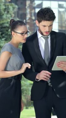 Kendine güvenen bir işadamı ve şık bir iş kadını, heyecan verici haberleri alan tabletleri tarafından büyülenen açık havada keyifli bir anı paylaşıyorlar. Gösterişli kıyafetleri, yemyeşil bir zemin ve modern mimari ile tamamlanmış.