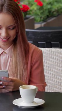 Pembe bir bluz giymiş ışıl ışıl genç bir kadın şık bir kafe masasında oturur, kahvesini yudumlarken akıllı telefonuyla eğlenir. Etrafı yemyeşil çiçeklerle çevrili olan manzara sıcaklığı ve canlı bir atmosferi yayar ve onu yakalar.