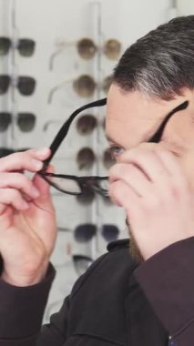 Moda gözlüklerini denerken mutlu, şık sakallı, neşeli, olgun bir adam şık çerçevelerle dolu canlı bir gözlük mağazasında başparmağını kaldırıyor. Bu dinamik sahne heyecan ve güven yayar.