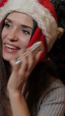 Noel Baba şapkalı neşeli beyaz bir kadın cep telefonuyla sohbet ederken ışıldayan süsler ve parlak ışıklarla süslenmiş çarpıcı bir Noel ağacı ile çevrili. Sıcak gülüşü ve rahat süveteri bayram neşesi saçıyor.