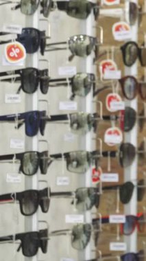 Dalgalı saçlı şık bir genç kadın, modern bir optometriste sergilenen bir dizi güneş gözlüğünü tutkuyla fotoğraflıyor. Kırsal tuğla duvarın sıcak renkleri canlı gözlüklerle çok güzel tezat oluşturuyor.