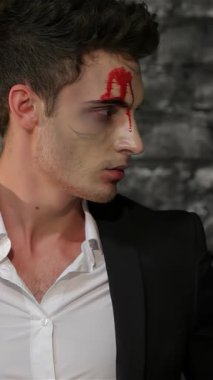 Gösterişli siyah takım elbiseli büyüleyici bir erkek vampir koyu renkli tuğla bir duvara karşı dramatik bir poz veriyor. Alnından kan kırmızısı bir aksan damlayan çarpıcı makyajı, Cadılar Bayramı 'nın ürkütücü cazibesini artırıyor, izleyicileri kendine çekiyor.