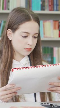 Uzun kahverengi saçlı kararlı bir genç kız kendini derslerine verirken elinde bir defter tutarak modern bir masada oturuyor. Renkli kitaplıklarla çevrili atmosfer akademik coşku ve merak yayıyor, mükemmel bir şekilde yakalıyor.