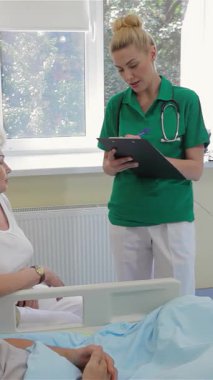 Parlak bir hastane koğuşunda, yeşil polo gömlekli şefkatli bir hemşire bir erkek hasta ve yaşlı karısıyla sıcak bir ilişki kurarak destekleyici bir atmosferi teşvik ediyor. Özenli tavrı ve güven veren gülüşü rahatlatıcı bir ortam yaratıyor.