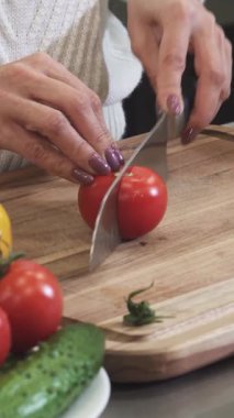 Yakın plan çekimlerde zarif manikürlü tırnakları olan bir kadın, bir dizi taze sebzeyle çevrili, kırsal bir tahta üzerinde parlak kırmızı bir domatesi ustalıkla dilimliyor. Bu canlı mutfak sahnesinde sıcaklık ve aşçılık tutkusu var.