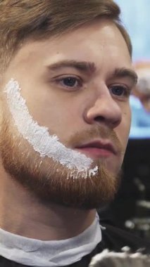 Şık bir berber dükkanında, yetenekli bir berber yakışıklı genç bir adamın sakalına ustalıkla tıraş kremi sürüp tımar deneyimini yükseltir. Kaliteli dekor ve sıcak ışıklandırmayla süslenmiş davetkar atmosfer mükemmel bir şekilde tamamlayıcı.