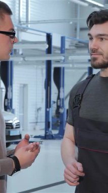 Gösterişli, modern bir atölyede, odaklanmış esmer bir tamirci, gözlük takan genç bir müşterisiyle araba tamirini tutkuyla tartışıyor. El sıkışmaları güven ve profesyonelliği sembolize ederek otomotiv hizmetindeki güçlü bağlantıyı vurguluyor.